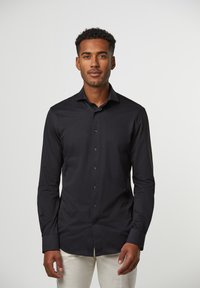 Zwarte langwerpige shirt met lange mouwen, gemaakt van gladde stof, met een knopenfront, puntige kraag en een aansluitend ontwerp. Gecombineerd met een lichte broek.