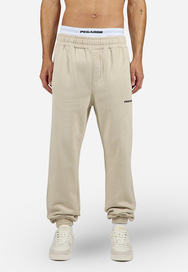 Beige joggebukser med elastisk linning, med en hvit logo og ribbede mansjetter. Laget av mykt materiale med en avslappet passform.