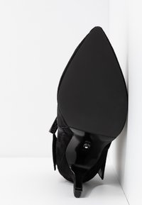 Chaussure à talon haut en daim noir à bout pointu, présentée semelle vers le haut sur un fond blanc en angle.
