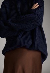 Tummansininen neulottu villapaita, jossa on ribbitekstuuri, kaapelitekniikka hihansuissa, löysä malli, käytetty ruskean hameen päällä.