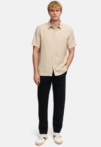 Homme blond debout portant une chemise beige à manches courtes boutonnée, un pantalon noir, et des baskets blanches avec semelles en gomme sur un fond clair uni.