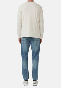 Camicia a maniche lunghe color crema abbinata a jeans azzurri chiari, con texture sbiadita e due tasche posteriori. Le scarpe sono bianche con accenti verdi.