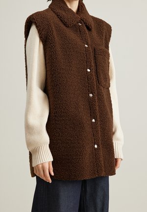 Mouwloos bruin sherpa-vest met drukknopen en borstzak, gedragen over een crème ribgebreide trui en donkere broek.