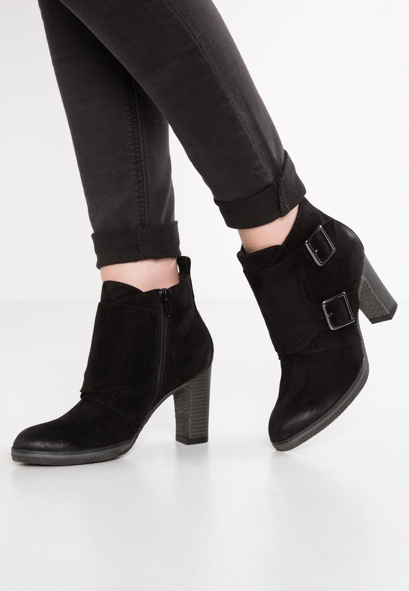 Bottines en daim noir à talons blocs, fermeture éclair latérale et double lanières à boucles, portées avec un jean noir retroussé sur un sol blanc.