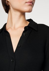 Haut en tricot noir avec un col, comportant une patte de boutonnage et un tissu texturé. Gros plan mettant en valeur les boutons et le design du col.