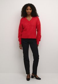Maglione rosso a maglia con scollo a V e maniche lunghe, abbinato a pantaloni neri slim-fit e tacchi neri lucidi. Tessuto strutturato con una vestibilità morbida.