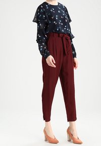 Blusa blu navy con motivo floreale, ruches sovrapposte, abbinata a pantaloni a vita alta color bordeaux e a scarpe con tacco a blocco traslucide.