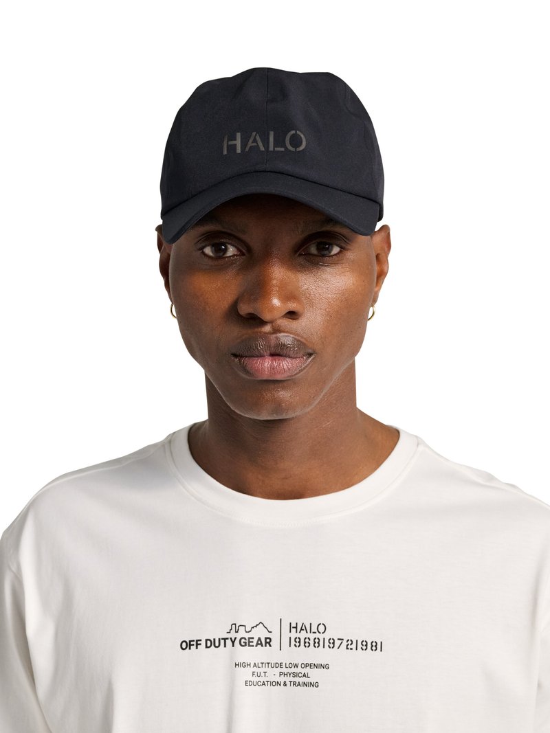 Jeune homme portant une casquette noire avec le texte "HALO" et un T-shirt blanc avec l'inscription "OFF DUTY GEAR" et un petit texte imprimé.