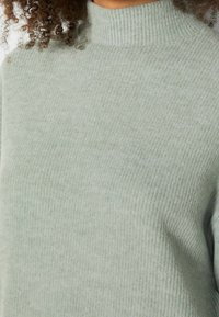 Personne portant un pull en tricot côtelé vert clair avec un col haut et des cheveux bouclés visibles aux épaules.