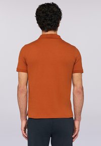 Rdzawoczerwona koszulka polo z krótkim rękawem, klasyczny kołnierzyk, gładka faktura i boki rozcięte u dołu. Widok z tyłu.
