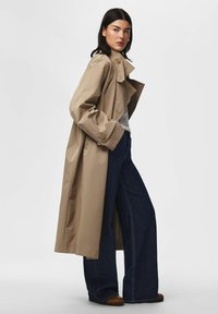 Femme aux cheveux lisses et foncés, portant un trench beige, un haut gris clair, un jean bleu foncé à jambes larges et des chaussures marron, debout de profil.