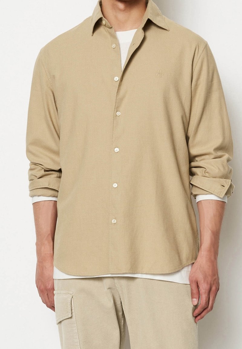 Homme portant une chemise beige à manches longues boutonnée par-dessus un sous-pull blanc, associée à un pantalon cargo beige, debout devant un fond blanc.