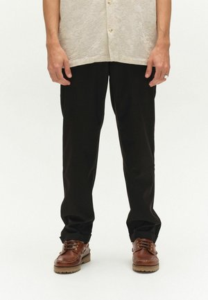 Mees, kes kannab musta lahti istuvat pükst, pruune nahast paeltega kingi ja helebeeži mustrilist särki ning seisab valgel taustal.