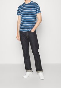 Blå och vit randig kortärmad t-shirt, matchad med mörka jeans och vita sneakers. T-shirten har en liten logotyp på bröstet.