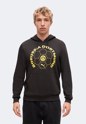 Junger Mann mit lockigem blondem Haar, der einen schwarzen Borussia Dortmund Hoodie mit gelbem Logo und Schriftzug trägt, vor einem schlichten Hintergrund stehend.