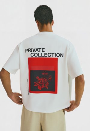 Mężczyzna noszący biały oversize t-shirt z wyraźnym napisem „PRIVATE COLLECTION” oraz czerwonym kwiatowym motywem na plecach, sparowany z beżowymi spodniami.