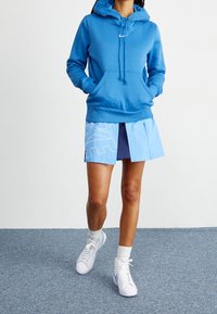 Blå hoodie med en ficka fram och dragsko på huvan, tillsammans med en delad ljusblå kjol med ett vitt geometriskt mönster. Vita sneakers.