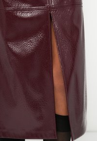 Φούστα από ψεύτικο δέρμα σε μπουρντό χρώμα με γυαλιστερό φινίρισμα, που διαθέτει πλαϊνή σχισμή. Επιφάνεια με υφή, διακοσμημένη με διακριτικά σχέδια και ραφές προσαρμοσμένες για καλύτερη εφαρμογή.