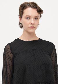 Junge Person mit kurzem, gewelltem Haar, die Hoop-Ohrringe und eine schwarze, durchsichtige Bluse mit Punkten trägt und direkt in die Kamera schaut.
