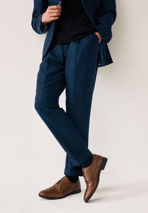 Moški v temno modrih hlačah in suknjiču z črno srajco, obut v rjave usnjene elegantne čevlje, stoji pred svetlim ozadjem.