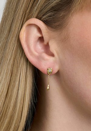 Primer plano de la oreja de una mujer con cabello rubio liso, que lleva un pendiente cuadrado de oro con una pequeña cadena y un colgante en forma de lágrima.