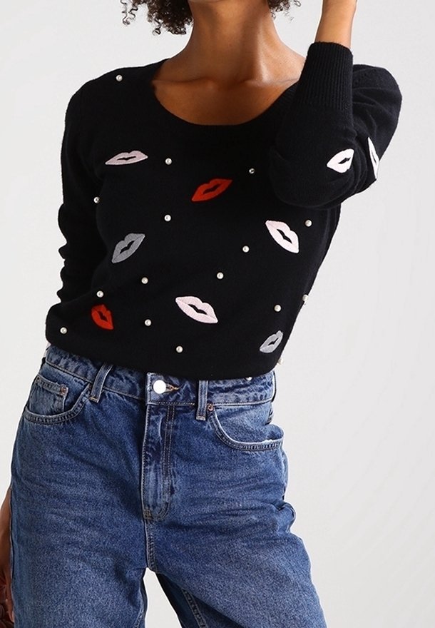 Pull noir avec des lèvres brodées en rouge, rose et gris, agrémenté de petites perles blanches, associé à un jean en denim bleu taille haute.