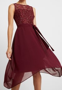 Ärmlös vinröd klänning med blommig spetsöverdel, genomskinlig lagerkjol och midjeband, bärs av en stående person mot en enkel bakgrund.