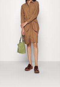 Abito marrone con bottoni e dettaglio annodato in vita; abbinato a una borsa in suede verde e zoccoli color bordeaux. Tessuto liscio e vestibilità sartoriale.