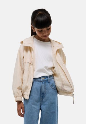 Veste beige clair avec fermeture éclair, capuche et poignets élastiques, portée sur un t-shirt blanc et un jean bleu taille haute ; doublure en mesh.