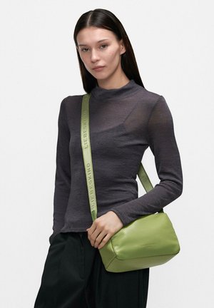 Kobieta o prostych ciemnych włosach nosi dopasowaną ciemnoszarą bluzkę z długim rękawem i trzyma jasnooliwkową torbę na ramię z szerokim paskiem.