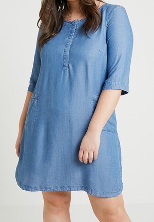 Kvinde iført en knælængde, lyseblå denimkjole med trekvartærmer og forlommer mod en ensfarvet baggrund.