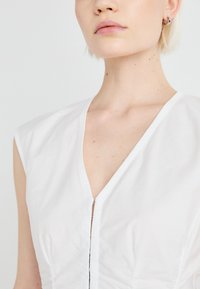 Blouse blanche sans manches avec un décolleté en V profond et une fermeture à l'avant. Texture lisse, design minimal et détails de couture subtils.