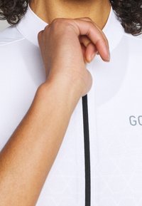 Biała koszulka kolarska z lekkiego materiału, z wysokim kołnierzem, zamkiem błyskawicznym i czarną linią ozdobną z przodu.