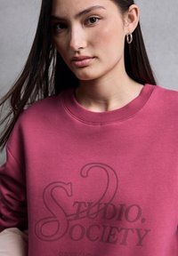 Felpa rosa con scollatura a costine e polsini. Presenta una stampa grafica scura con la scritta "STUDIO. SOCIETY" e uno slogan creativo sottostante.