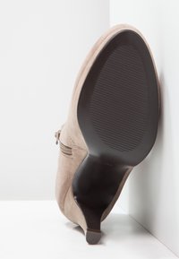 Botín de tacón alto de ante beige con cremallera lateral, suela negra y punta afilada, apoyado contra una pared blanca en la esquina.