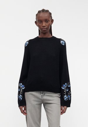 Femme portant un pull noir avec une broderie florale bleue sur les épaules et les manches, associé à un jean gris clair, sur fond blanc.