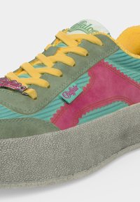 Gros plan d'une basket colorée avec des lacets jaunes, des panneaux en daim verts et roses, une semelle grise texturée et une étiquette logo près des lacets et du côté.