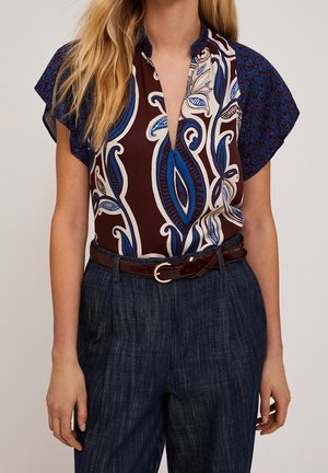 Kvinde iført en ærmeløs top med blå og brune blomsterprint, stoppet ned i højtaljede mørkeblå bukser og accessoriseret med et mørkebrunt bælte.