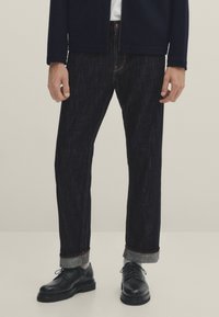 Tummansiniset farkut suoralla mallilla, joissa on käännetyt lahkeet, kontrastivärinen tikkaus ja nap fly, yhdistettynä mustiin nahkakenkiin.