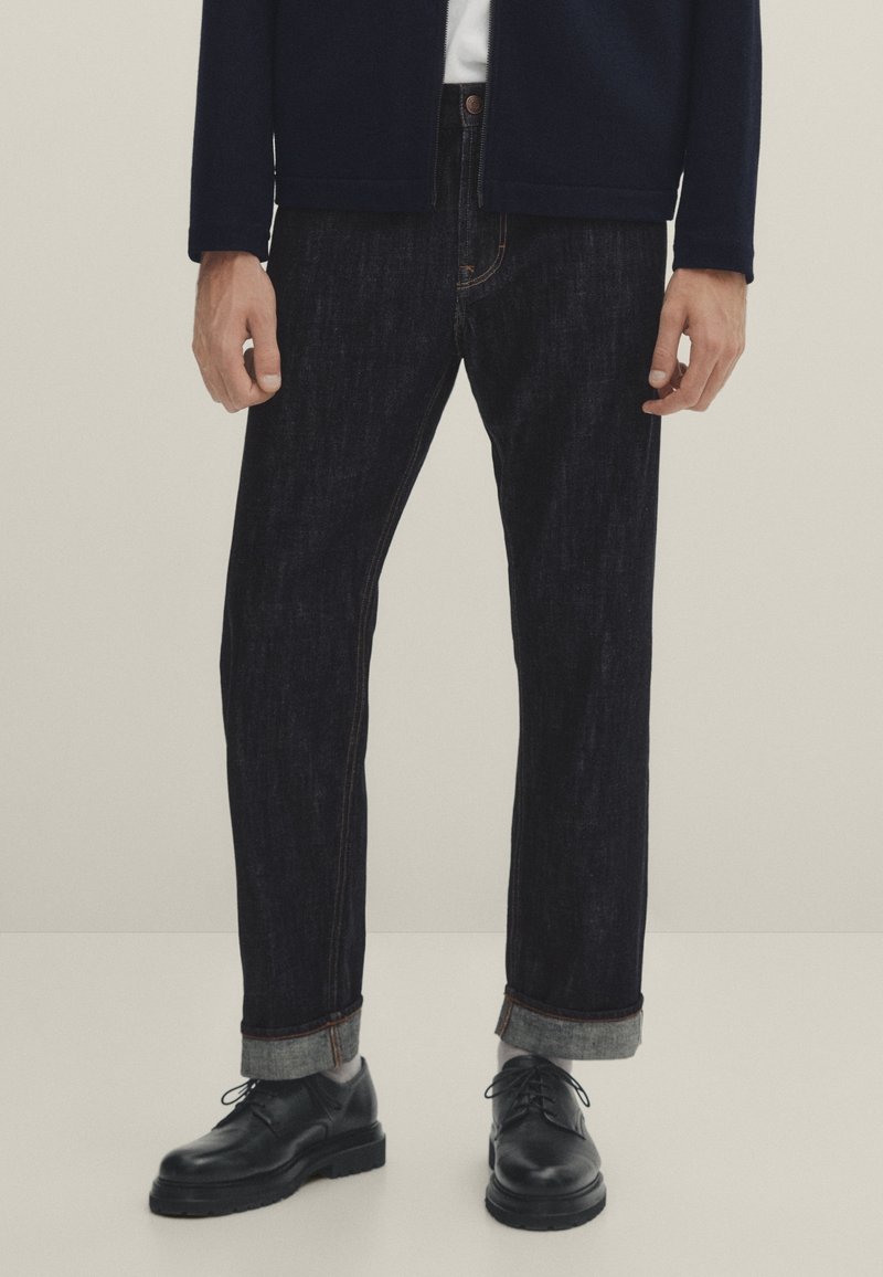 Tummansiniset farkut suoralla mallilla, joissa on käännetyt lahkeet, kontrastivärinen tikkaus ja nap fly, yhdistettynä mustiin nahkakenkiin.