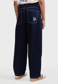 Pantalons en denim noir à jambes larges avec des coutures blanches et des poches à patte. Présente un logo sur la poche droite. Portés avec des baskets claires.