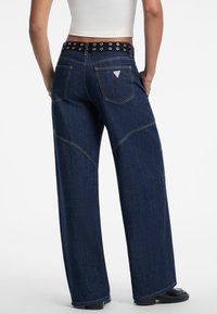 Wijd vallende donkerblauwe spijkerbroek met een hoge tailleband, voorzien van een zwarte riem met zilveren oogjes en achterzakken. Kenmerkende stiknaaddetails.