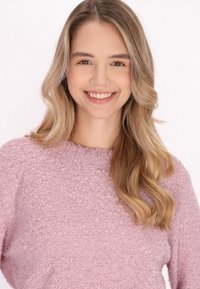 Pulover texturat roz deschis, cu guler rotund. Modelul are părul lung și ondulat. Trăsături faciale blânde, cu fard de pleoape portocaliu subtil. Fond alb.