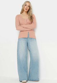 Ljusrosa ribbad cardigan med rundad halsringning och knappstängning, kombinerad med vida ljusblå jeans och beige skor.