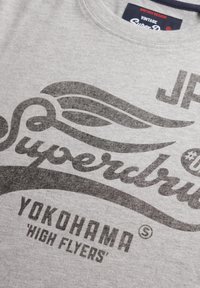 T-shirt en coton gris avec du texte imprimé noir, incluant "Superdry" et "Yokohama High Flyers", agrémenté d'accents graphiques d'ailes.