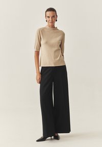 Beżowy top z krótkim rękawem i wysokim dekoltem, połączony z czarnymi spodniami o szerokich nogawkach oraz czarnymi butami na szpilce. Gładka faktura, minimalistyczny design.