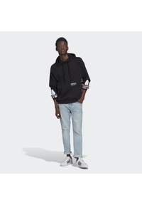 Felpa nera con cappuccio e logo bianco, con maniche decorate da strisce rosse e blu. Indossata con jeans azzurri chiari e sneakers bianche.