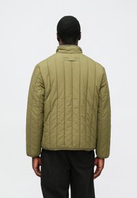 Casaco acolchoado verde-oliva com design acolchoado vertical, gola alta e duas abas na parte de trás. O tecido aparenta ser liso com acabamento mate.