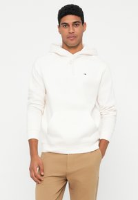 Muž v bielom mikine s kapucňou a béžových nohaviciach, stojaci s jednou rukou v vrecku na pozadí s bielou farbou.