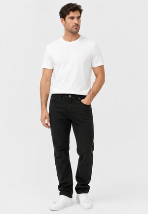 Man staat, draagt een effen wit T-shirt, zwarte spijkerbroek en witte sneakers, met één hand in zijn zak, kijkt naar links.